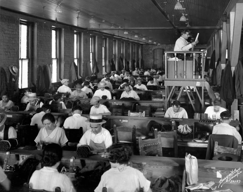 The interior of a large Tampa cigar factory: long rows of workers rolling cigars at benches, with the lector reading aloud from a raised platform on the right.