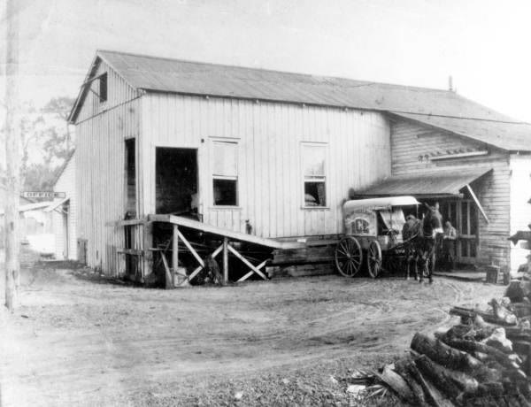 A horse-drawn ice wagon with a canvas cover stands beside a wooden ice house; a man and a horse wait in the dirt yard.
