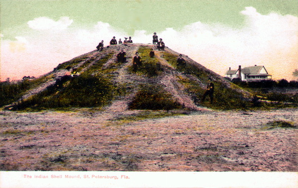 A hand-tinted circa-1900 view of a large Native American shell mound on the Tampa Bay shore, several people seated on its grassy slope, a house beside it.