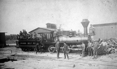 A small early steam locomotive of the narrow-gauge Orange Belt Railway on the tracks, several men in hats standing on and beside it.