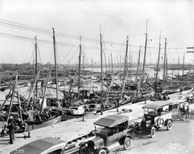 A long row of masted sponge boats moored along a dock, with a line of early automobiles and a horse-drawn wagon parked beside them and men sitting on the dock.